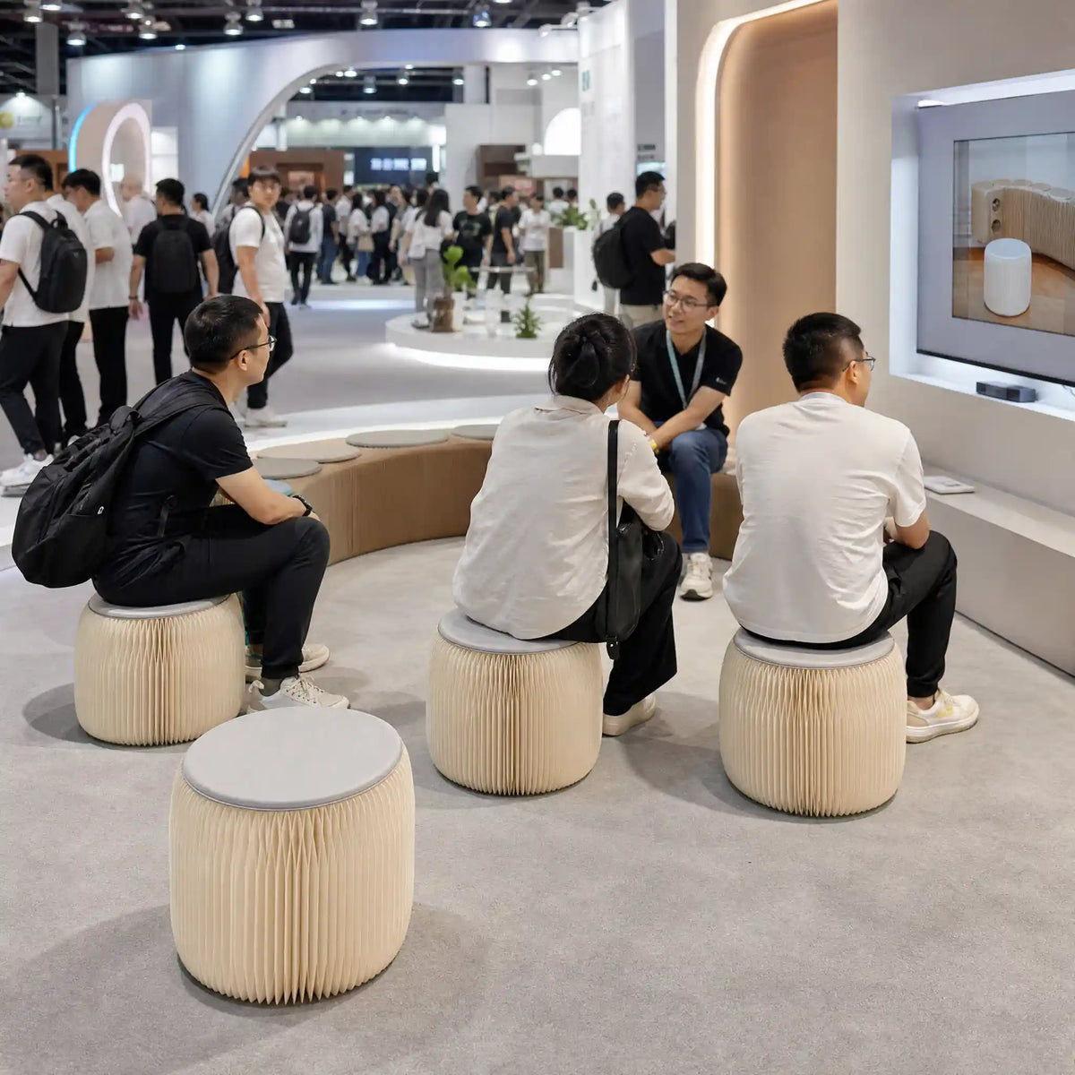 Attendees sitting on foldable paper stools around a display at a trade show booth.