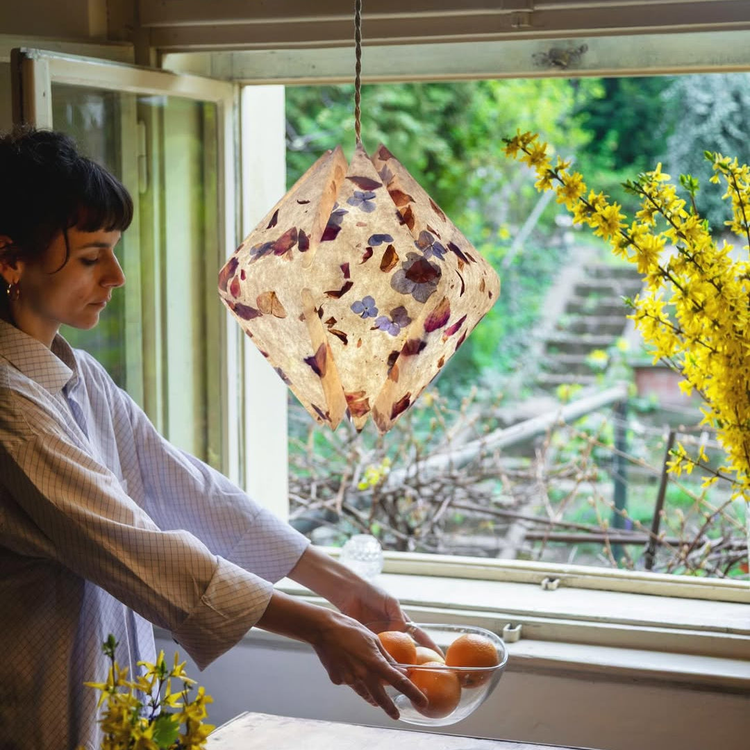 Woman adjusting botanical print origami paper pendant lampshade near window with dried flowers